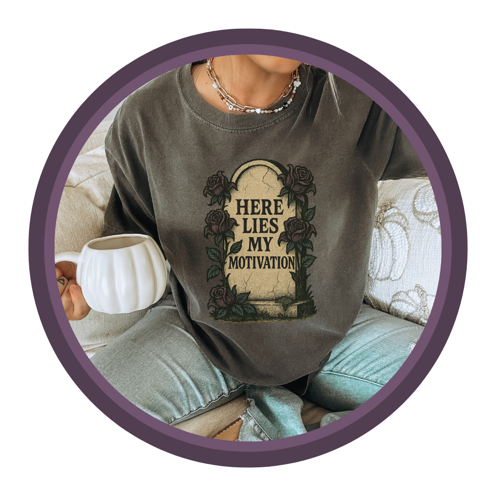 photo of a woman sitting on a couch holding a white pumpkin mug wearing a t-shirt that says "Here Lies My Motivation" on a tombstone.