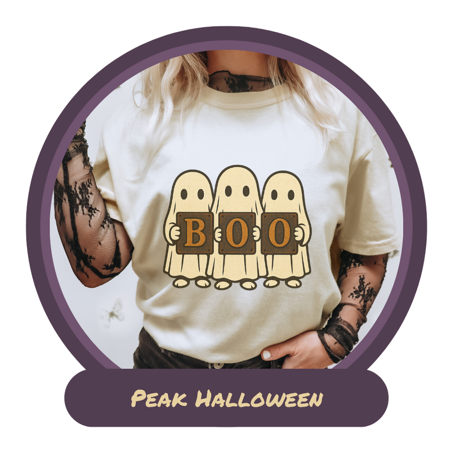 blonde woman wearing an ivory t-shirt over a black lace long sleeve shirt. Her t-shirt has 3 cute sheet ghosts holding up metal letters that spell out Boo