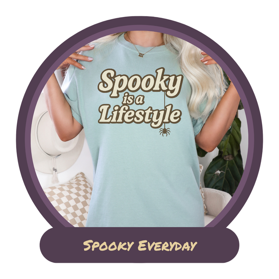 Blonde woman in a bay colored t-shirt that says Spooky is a Lifestyle in bold text with a small brown spider hanging from the words